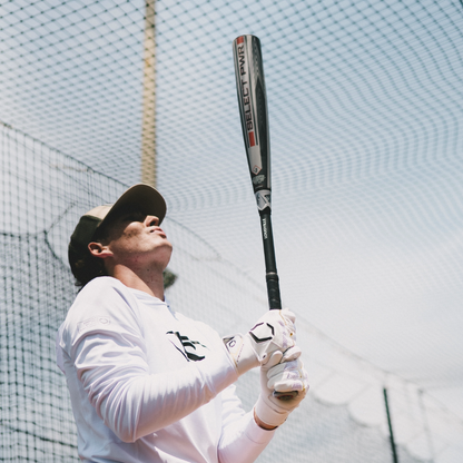 A baseball player in a white long-sleeve shirt and cap holds the Louisville Slugger 2026 Select PWR (-3) BBCOR Baseball Bat (WBL4115010) vertically, looking up with focus, standing before a netted fence under a clear sky, ready to practice or play.