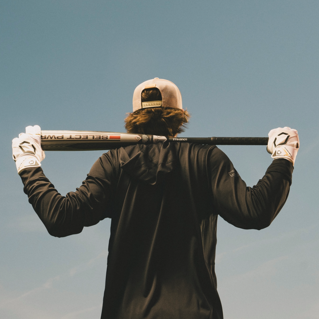 A person in a baseball cap, black long-sleeve shirt, and white gloves holds the Louisville Slugger 2026 Select PWR (-3) BBCOR Baseball Bat (WBL4115010) across their shoulders while facing away against a blue sky.