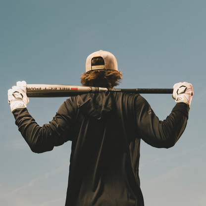 A person in a baseball cap, black long-sleeve shirt, and white gloves holds the Louisville Slugger 2026 Select PWR (-3) BBCOR Baseball Bat (WBL4115010) across their shoulders while facing away against a blue sky.