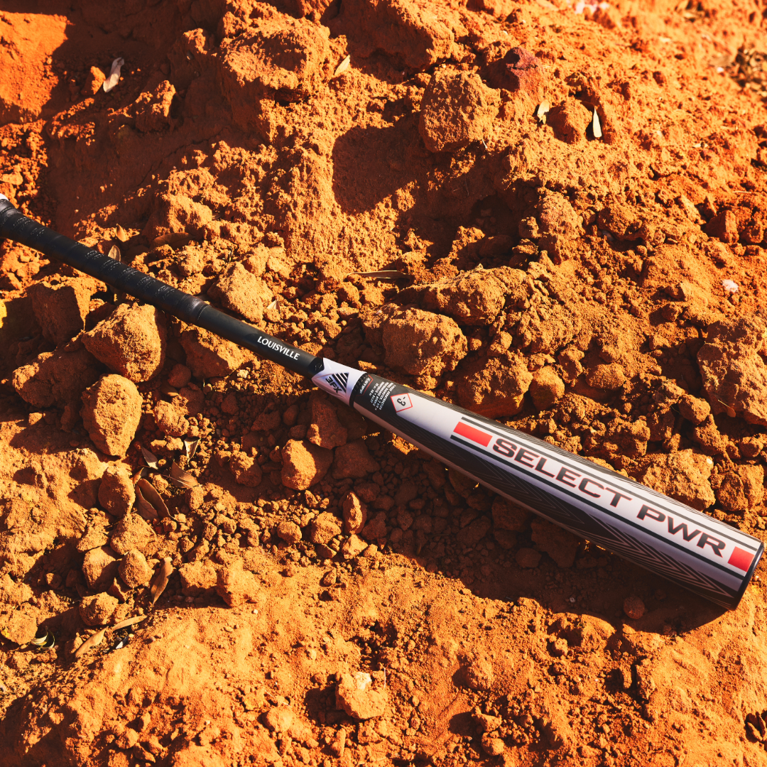 The 2026 Louisville Slugger Select PWR (-3) BBCOR Baseball Bat (WBL4115010), in sleek silver, black, and red, rests on bright orange dirt amid sunlit soil mounds—ideal for power hitters ready to make an impact.