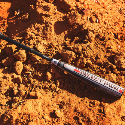 The 2026 Louisville Slugger Select PWR (-3) BBCOR Baseball Bat (WBL4115010), in sleek silver, black, and red, rests on bright orange dirt amid sunlit soil mounds—ideal for power hitters ready to make an impact.