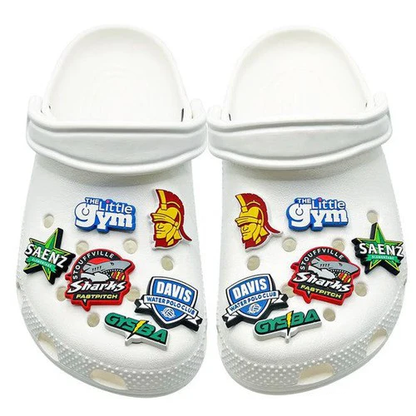 A pair of white Crocs decorated with DSG Apparel’s NSA Softball 3D Rubber Clog Charm and colorful jibbitz, featuring The Little Gym, a Trojan helmet, softball designs, and sports team badges.