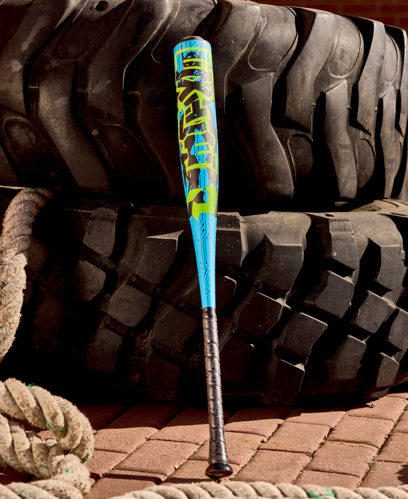 The DeMarini 2027 Exile (-5) USSSA Baseball Bat (WBD2716010), featuring a blue and yellow X14 Alloy 2 3/4" barrel and black grip, leans on large black tractor tires with thick white ropes coiled nearby on a brick surface.