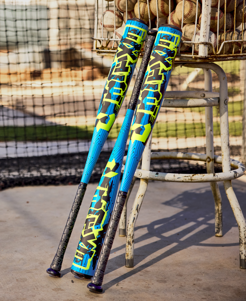Two vibrant DeMarini 2027 Exile (-5) 2 3/4" USSSA Baseball Bats (WBD2716010) with blue, yellow, and black designs lean on a basket of baseballs near a stool and cage, featuring X14 Alloy construction on a concrete floor.