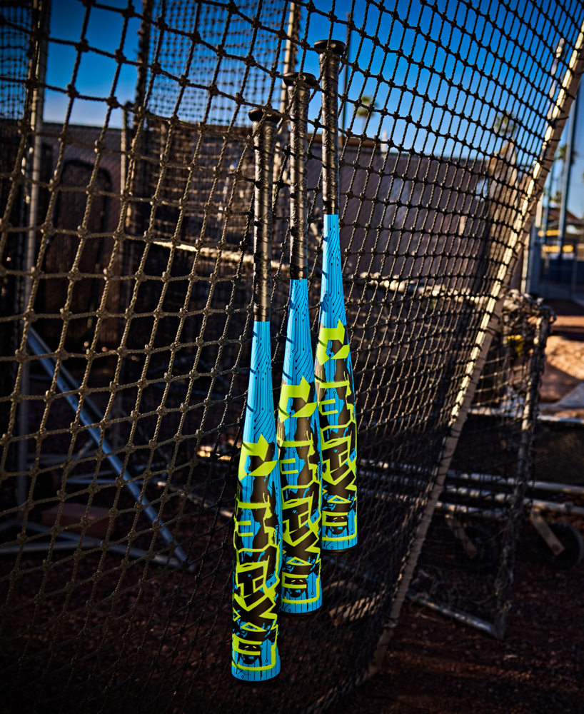 Three DeMarini 2027 Exile (-5) 2 3/4" USSSA Baseball Bats (WBD2716010), each with a one-piece design in blue and yellow, hang on batting cage netting with sports gear and fence visible beneath a clear sky.