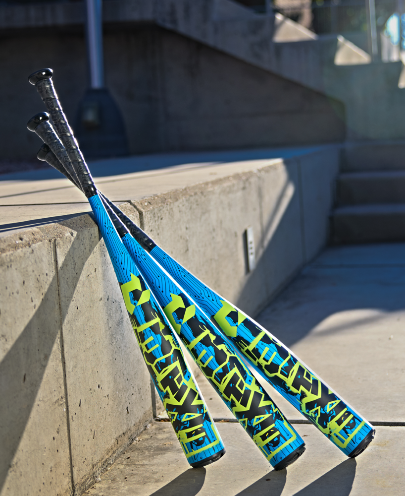 Three DeMarini 2027 Exile (-5) USSSA baseball bats with blue and yellow barrels (2 3/4") and black handles rest against a concrete wall outdoors, sunlight casting shadows on the ground and stairs behind them.