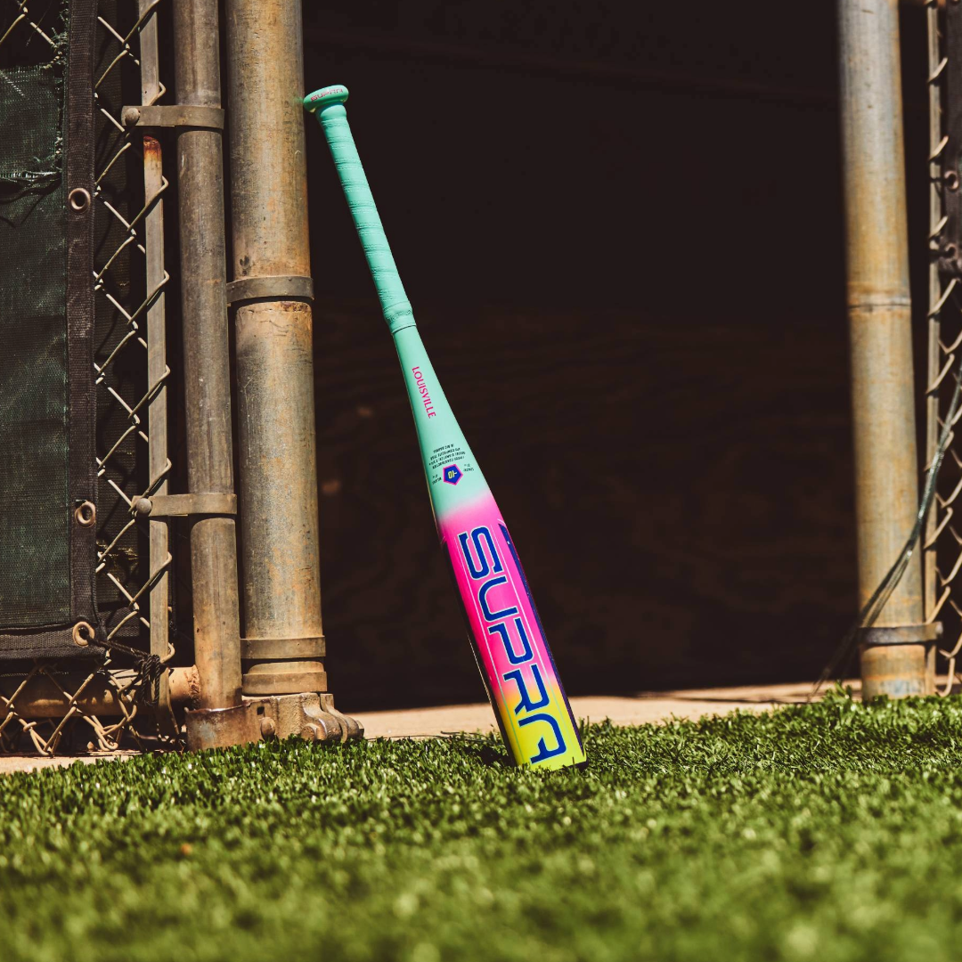 A 2027 Louisville Slugger Supra JBB (-10) USSSA Baseball Bat (WBL4265010) with “SUPRA” on the barrel leans against a metal gate on turf near a dugout entrance.