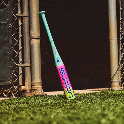 A 2027 Louisville Slugger Supra JBB (-10) USSSA Baseball Bat (WBL4265010) with “SUPRA” on the barrel leans against a metal gate on turf near a dugout entrance.
