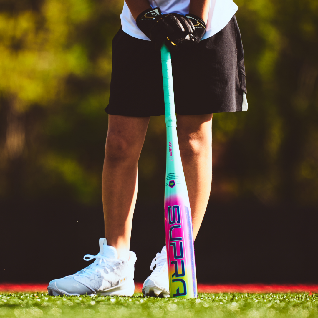 A child in white sneakers and a black skirt stands on a field, holding the 2027 Louisville Slugger Supra JBB (-10) USSSA Baseball Bat vertically, displaying the bold "SUPRA" lettering.