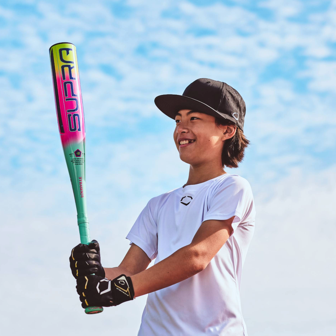 A smiling young person in a white shirt and black cap holds the 2027 Louisville Slugger Supra JBB (-10) USSSA Baseball Bat outdoors, under a bright blue sky with scattered clouds.