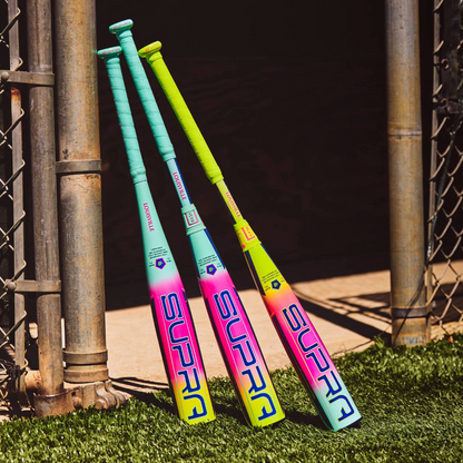 Three colorful Louisville Slugger Supra (-10) 2 5/8" USA Baseball Bats (WBL4258010) lean against a metal fence on artificial turf, showing off bright pink, yellow, blue, and green hues in the sunlight.