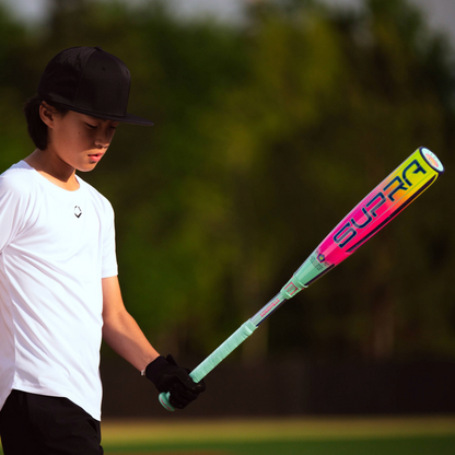 A young person in a white shirt and black cap stands on a baseball field holding the 2027 Louisville Slugger Supra (-10) 2 3/4" USSSA Baseball Bat (WBL4251010), with trees and blurred greenery in the background.