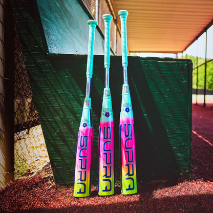 Three colorful Louisville Slugger baseball bats, including the 2027 Supra (-5) 2 3/4" USSSA Baseball Bat (WBL4253010), stand upright against a green wooden bench on a field, with a chain-link fence and dugout in the background.