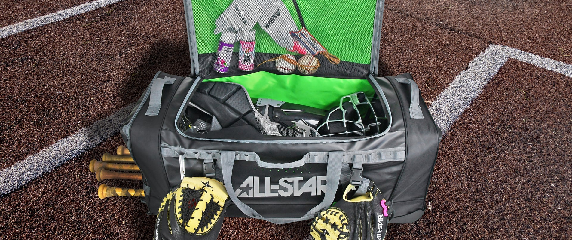 A large black and green All-Star sports bag is open on a baseball field, revealing gloves, a batting helmet, softballs, bats, and various gear inside and in the front pocket. Two yellow catchers mitts hang from the handles.