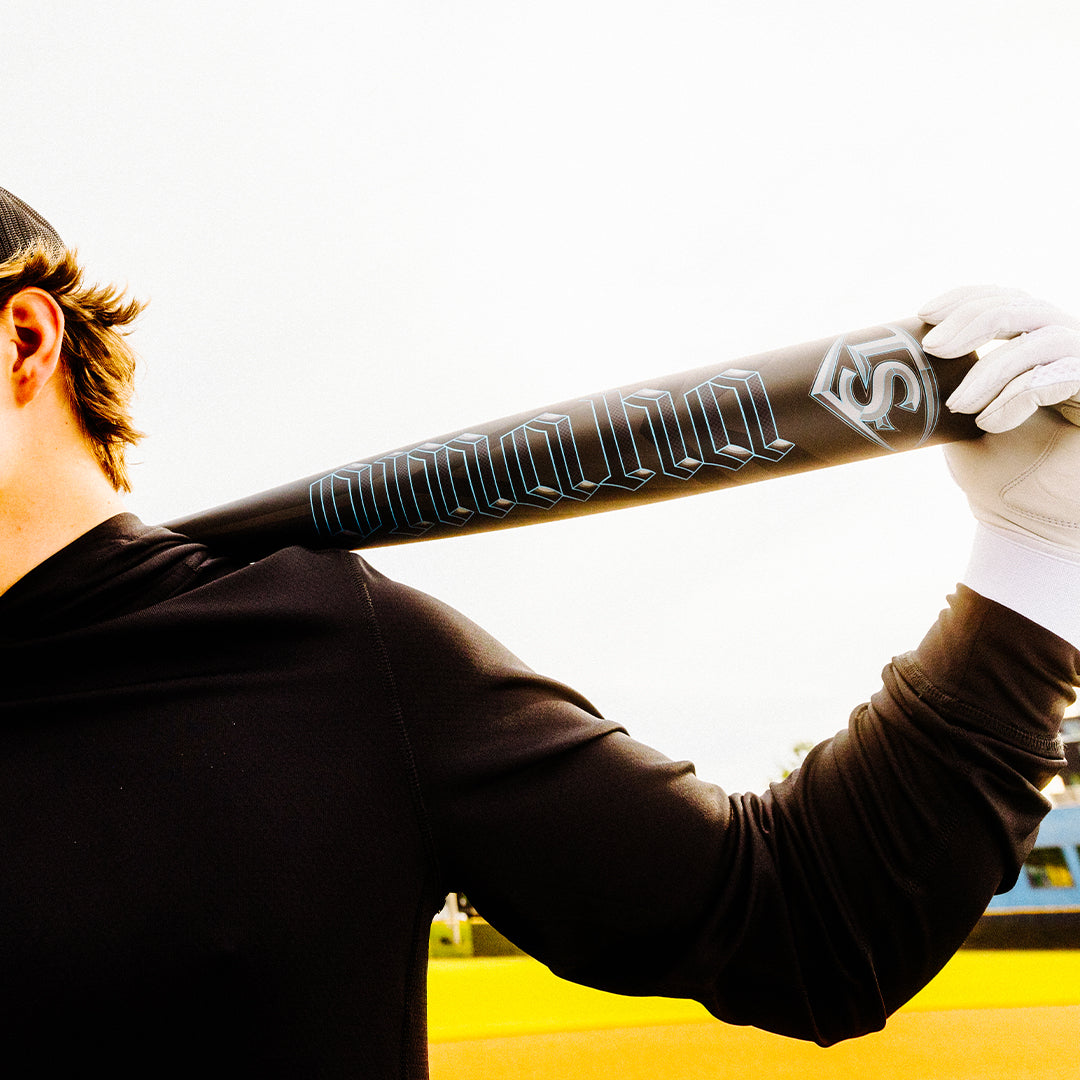 A baseball player in a black long-sleeve shirt and white glove holds a Louisville Slugger 2025 Omaha (-3) BBCOR Baseball Bat (WBL2984010) with silver lettering over their shoulder on the field, face partially out of frame.