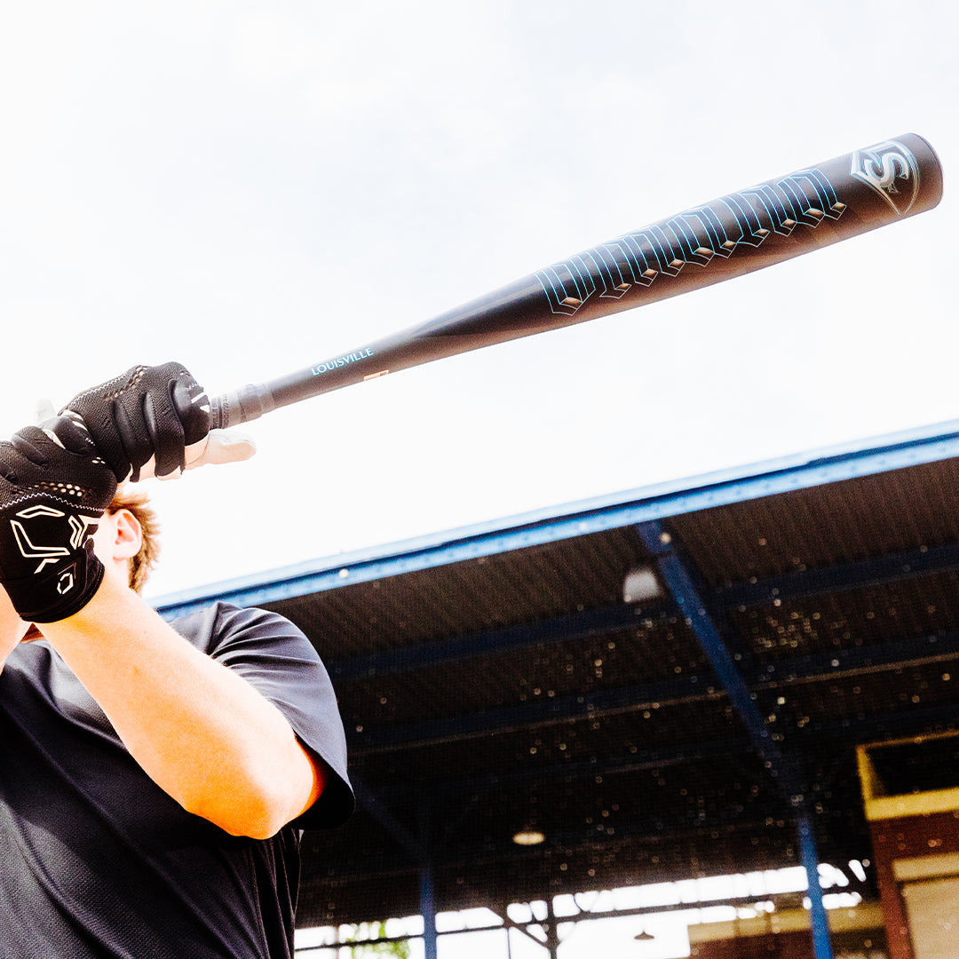 Wearing black gloves and a black shirt, a person prepares to swing the 2025 Louisville Slugger Omaha (-3) BBCOR Baseball Bat (WBL2984010) at an outdoor batting facility with a metal roof in the background.
