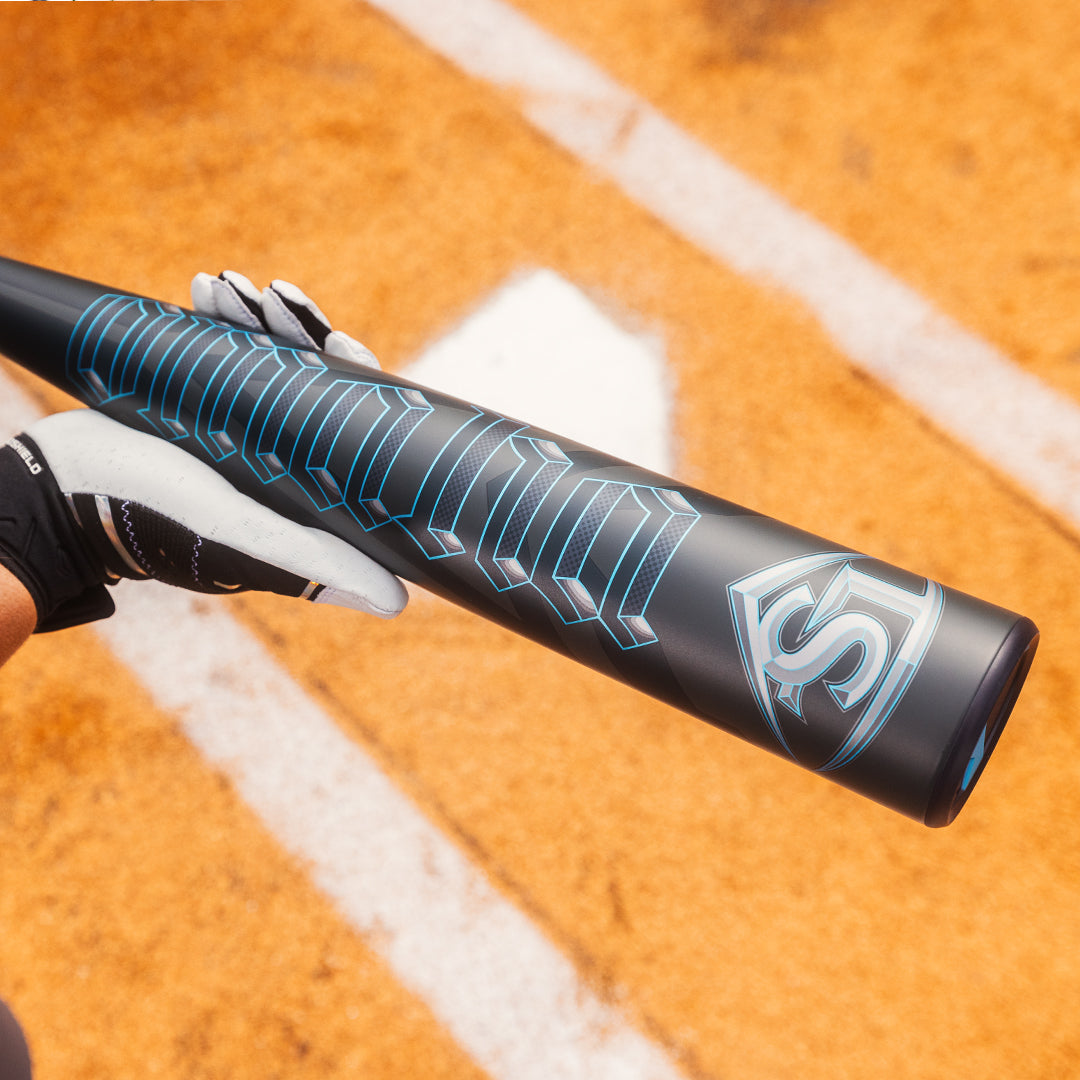 A person in a black and white glove holds the Louisville Slugger 2025 Omaha (-3) BBCOR Baseball Bat (WBL2984010), featuring blue and silver accents, above home plate on an orange-brown baseball field marked with white chalk lines.