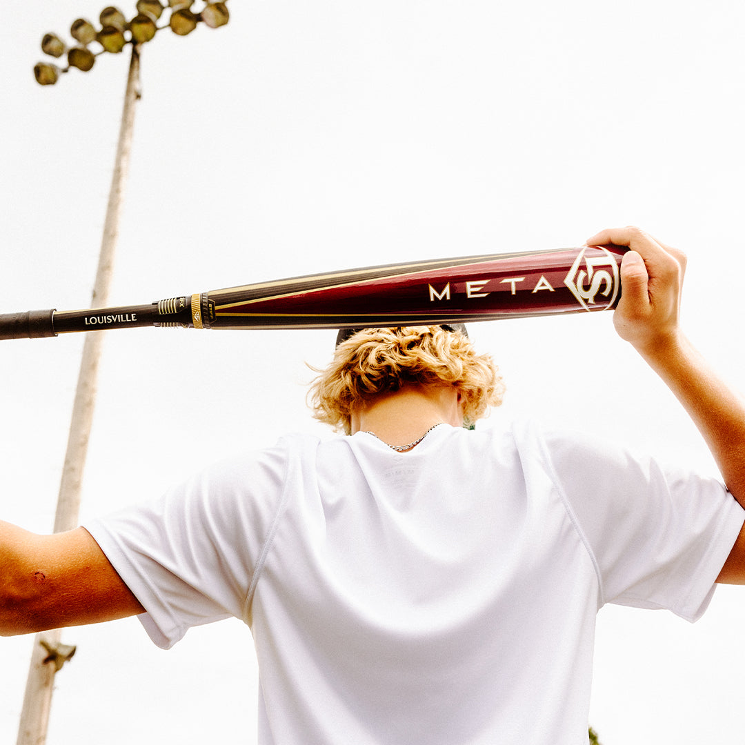 A person with blond hair in a white shirt holds a maroon 2025 Louisville Slugger Meta (-3) BBCOR Baseball Bat (USED) behind their neck, standing outdoors under bright lights against a pale sky.