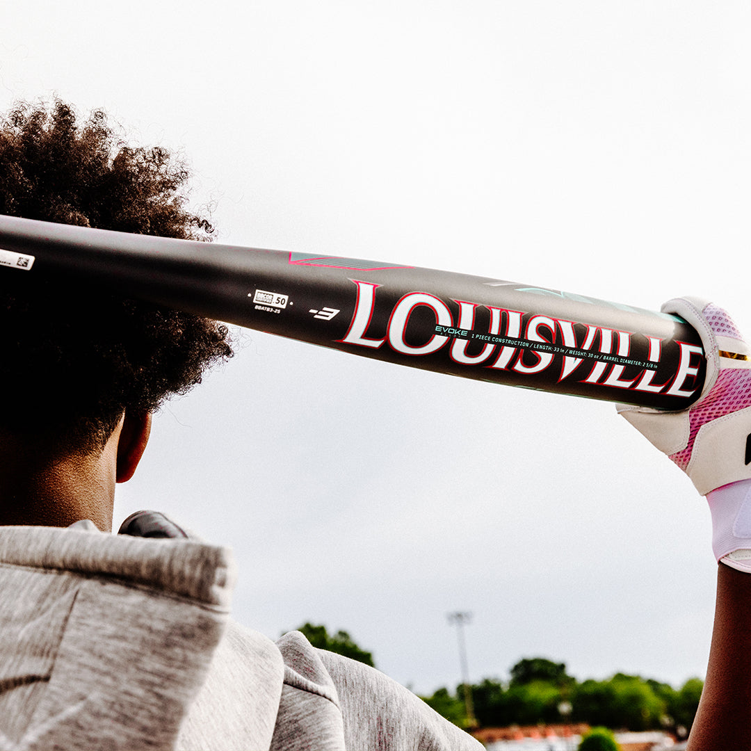 A person holds a black 2025 Louisville Slugger Atlas (-3) BBCOR Baseball Bat (WBL2968010 DEMO) over their shoulder, wearing a gray hoodie and white-and-pink glove, with trees and a cloudy sky in the background.
