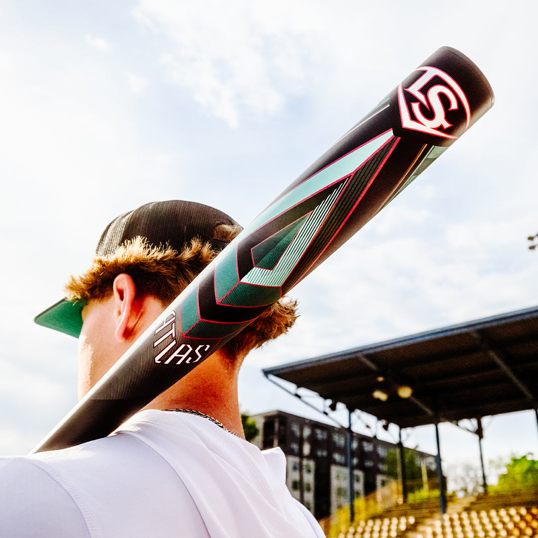 A baseball player with curly hair and a black cap holds the 2025 Louisville Slugger Atlas (-3) BBCOR Baseball Bat: WBL2968010 (DEMO) over his shoulder near bleachers at an outdoor baseball field on a sunny day.