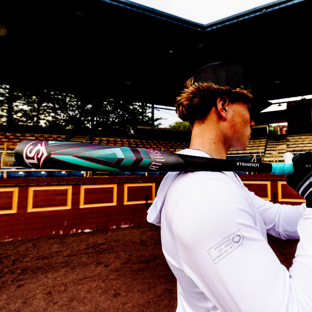 A baseball player in a white hoodie and black cap holds the Louisville Slugger 2025 Atlas (-3) BBCOR Baseball Bat (WBL2968010 DEMO) over his shoulder in an empty stadium with red and yellow walls.