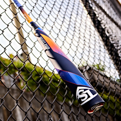 A 2025 Louisville Slugger Select PWR (-3) BBCOR Baseball Bat (DEMO) with blue, orange, and purple accents leans against a chain-link fence outdoors, sunlight and greenery in the background.