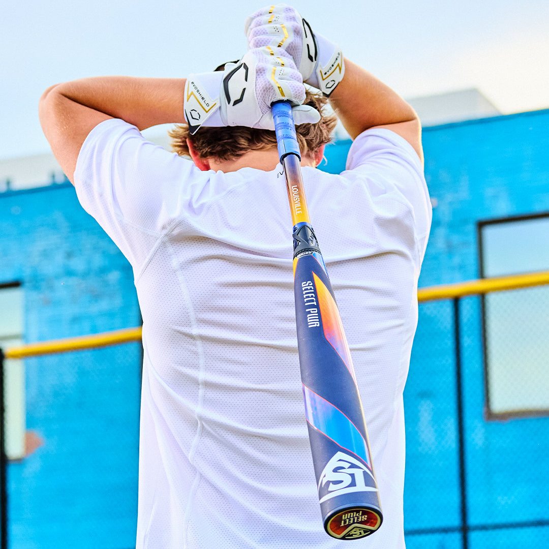 A person in a white shirt and gloves holds the 2025 Louisville Slugger Select PWR (-3) BBCOR Baseball Bat (WBL2967010 DEMO) behind their neck, viewed from the back, with a blue wall and yellow fence visible under a clear sky.