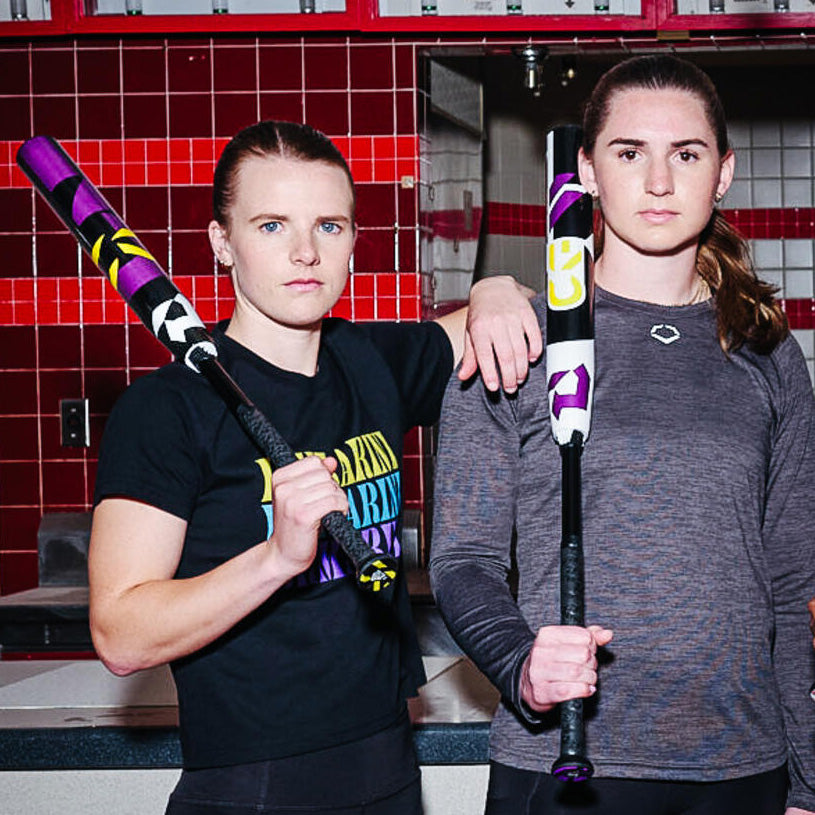 Two women stand indoors in front of red and white tiled walls, each holding a colorful softball bat. One woman wears a black shirt and the other a long-sleeve gray shirt, both looking confidently at the camera.
