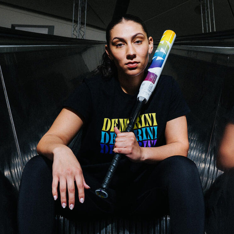 A woman sits on an escalator holding a colorful baseball bat over her shoulder, wearing a black t-shirt with bold multicolored text and black pants, looking confidently at the camera.