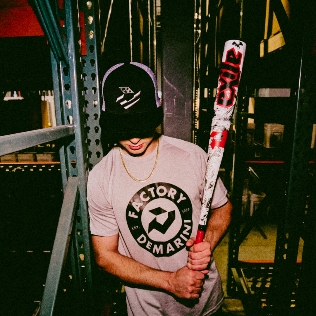 Wearing a black hat and Factory Demarini t-shirt, a person stands indoors among metal shelves, holding a white and red 2026 DeMarini Exile (-3) BBCOR Baseball Bat (WBD2589010) by DeMarini, their face partially hidden by the hat.
