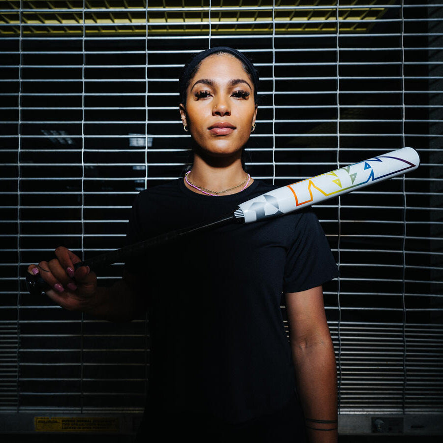 A woman in a black shirt stands confidently in front of a metal grate, holding a colorful baseball bat across her shoulders and looking directly at the camera.