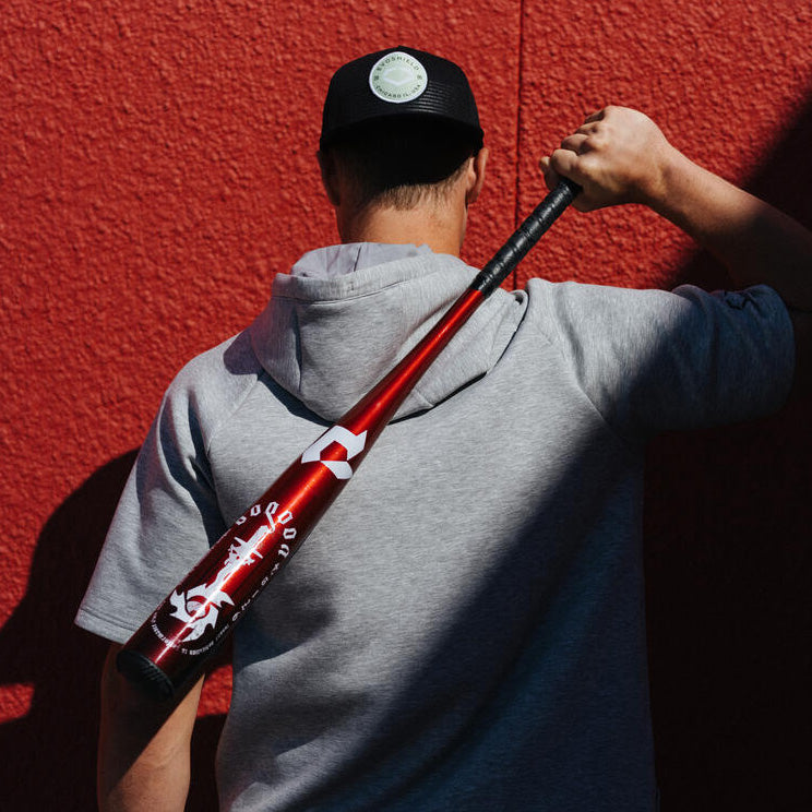 A person in a gray hoodie and black cap stands with their back to the camera, holding a red baseball bat over their shoulder, against a textured red wall.