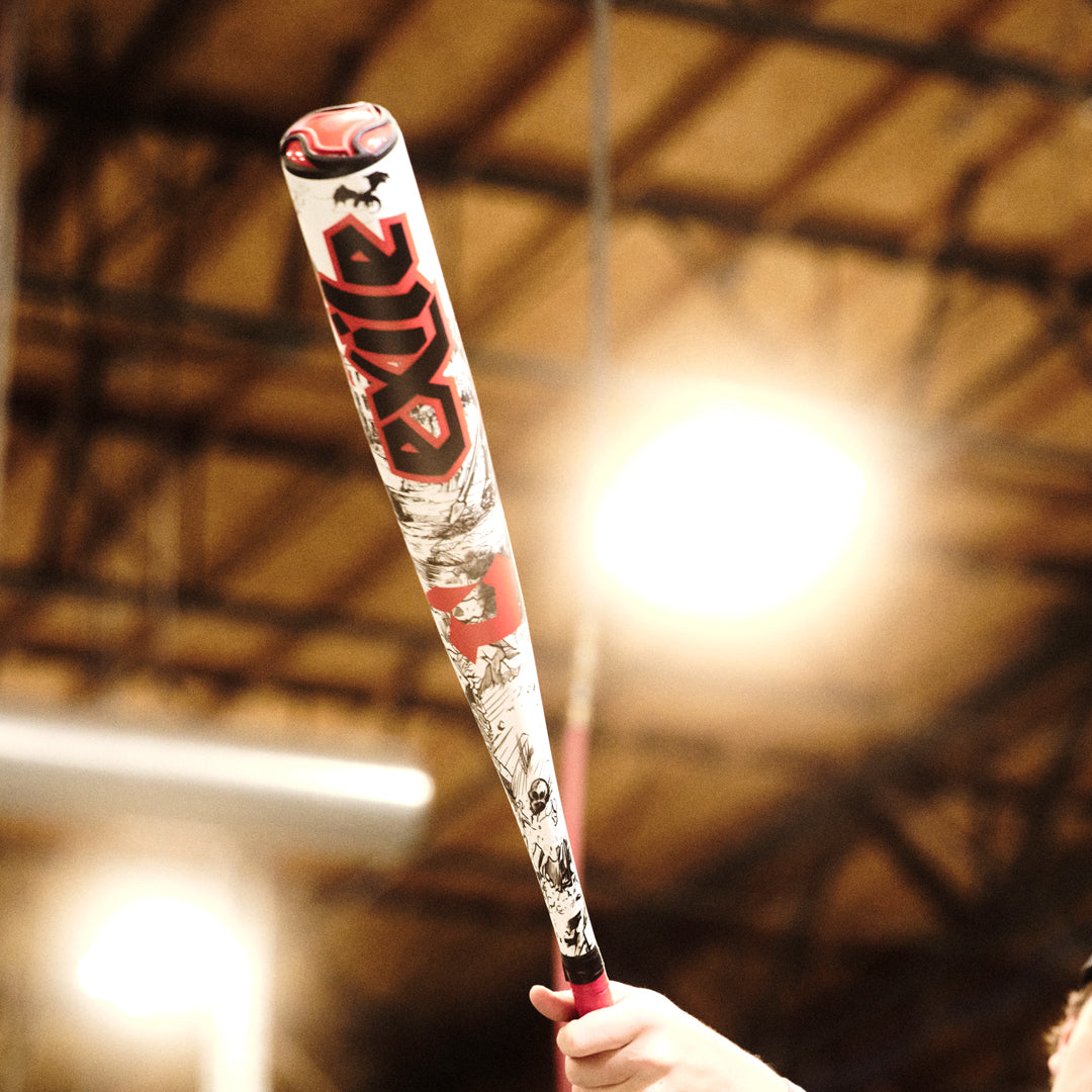 A person holds up the 2026 DeMarini Exile (-3) BBCOR Baseball Bat (WBD2589010) with red and black graphics, under bright indoor lights.