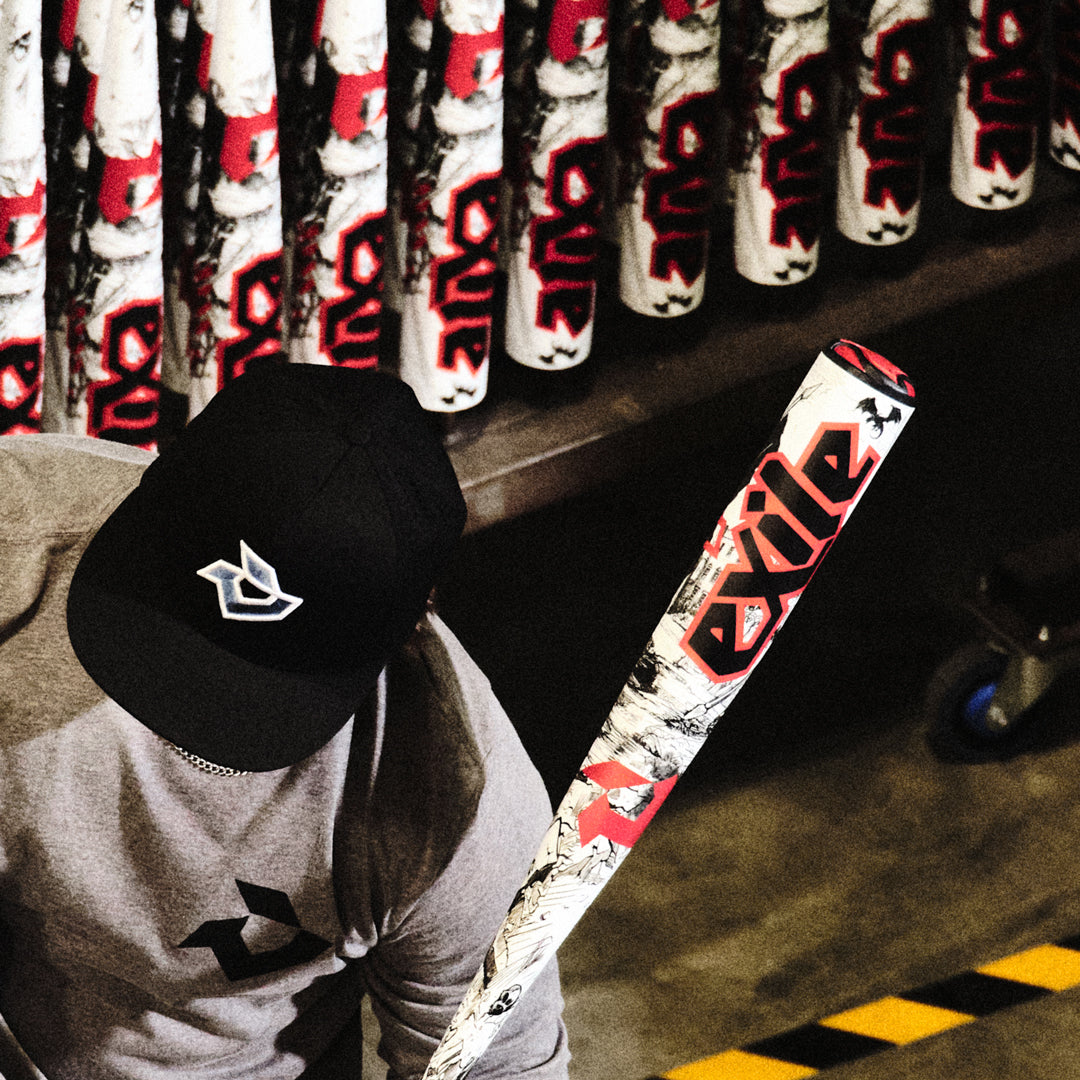 A person in a black cap and grey logo shirt examines a 2026 DeMarini Exile (-3) BBCOR Baseball Bat: WBD2589010, with numerous similar DeMarini bats standing upright in the background.