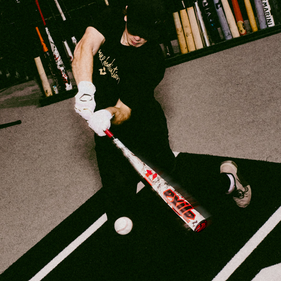 A baseball player in black swings a DeMarini 2026 Exile (-3) BBCOR Baseball Bat (WBD2589010) at a ball indoors, surrounded by upright bats in the background.