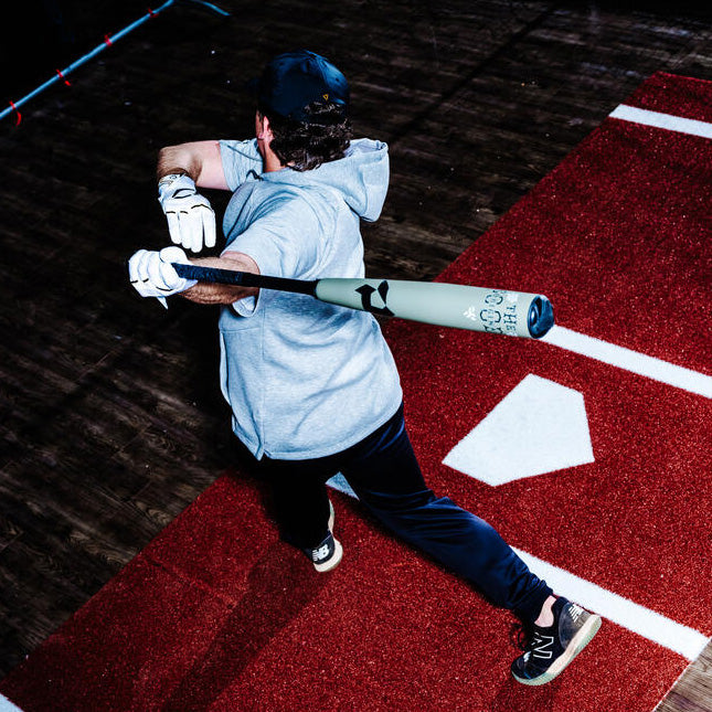 A baseball player wearing a gray hoodie and dark pants swings a bat near home plate on a red artificial turf field, seen from above.