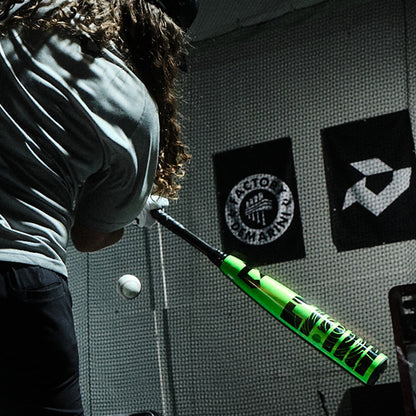 A person with long hair swings a neon green DeMarini 2026 ZEN (-8) 2 3/4" USSSA Baseball Bat (WBD2592010) at an incoming baseball in an indoor batting cage, black-and-white banners hanging in the background.