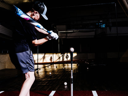 A young player in a cap and gloves swings a 2025 DeMarini ZEN (-5) 2 3/4" USSSA Baseball Bat (USED) at a ball on a tee in an indoor cage, with scattered baseballs littering the dark floor.