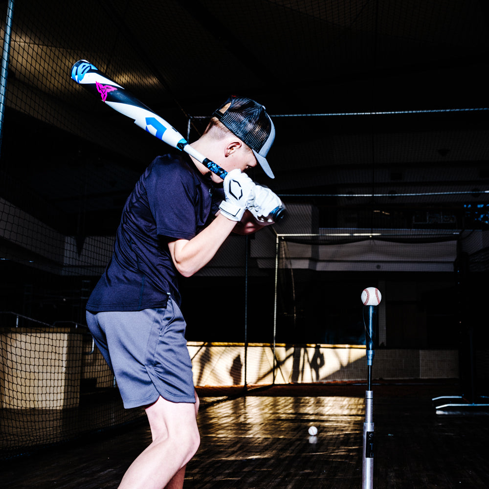 A young baseball player, dressed in athletic gear, gets ready to swing a 2025 DeMarini ZEN (-5) 2 3/4" USSSA Baseball Bat (USED) at a ball on a tee inside an indoor training facility.