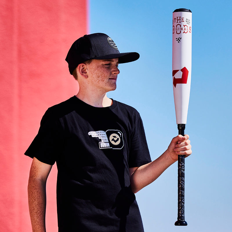 A young man in a black hat and t-shirt holds a white baseball bat with black and red designs, standing against a background split into red and blue.