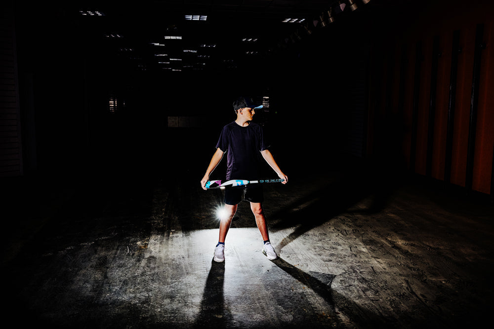 Wearing a cap, t-shirt, and shorts, a person stands in a dark indoor space holding a colorful 2025 DeMarini ZEN (-10) 2 3/4" USSSA Baseball Bat (WBD2532010, USED), with light behind them casting dramatic shadows.