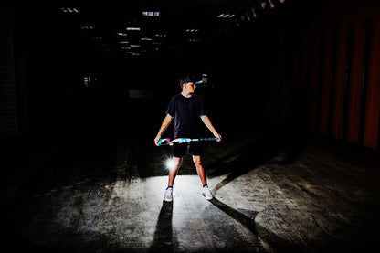 Wearing a cap, t-shirt, and shorts, a person stands in a dark indoor space holding a colorful 2025 DeMarini ZEN (-10) 2 3/4" USSSA Baseball Bat (WBD2532010, USED), with light behind them casting dramatic shadows.
