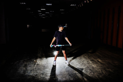 In a dark room, a person stands under a spotlight, gripping the 2025 DeMarini ZEN (-8) 2 3/4" USSSA Baseball Bat (WBD2533010). Wearing a cap, dark t-shirt, shorts, and white sneakers, they cast dramatic shadows and light on the floor.