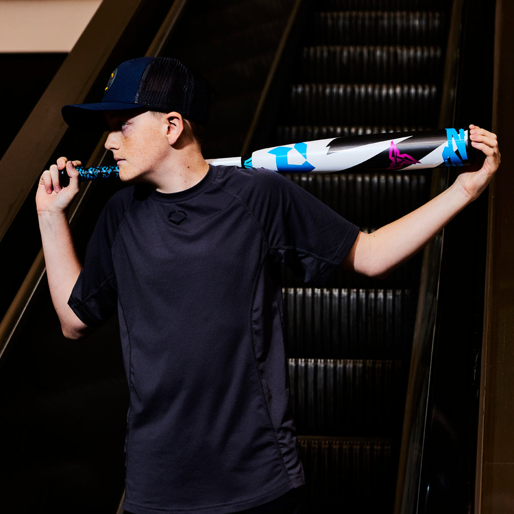 A boy in a dark shirt and cap stands before an escalator, holding a DeMarini 2025 ZEN (-5) 2 3/4" USSSA Baseball Bat (WBD2534010, USED) across his shoulders and glancing to the side.