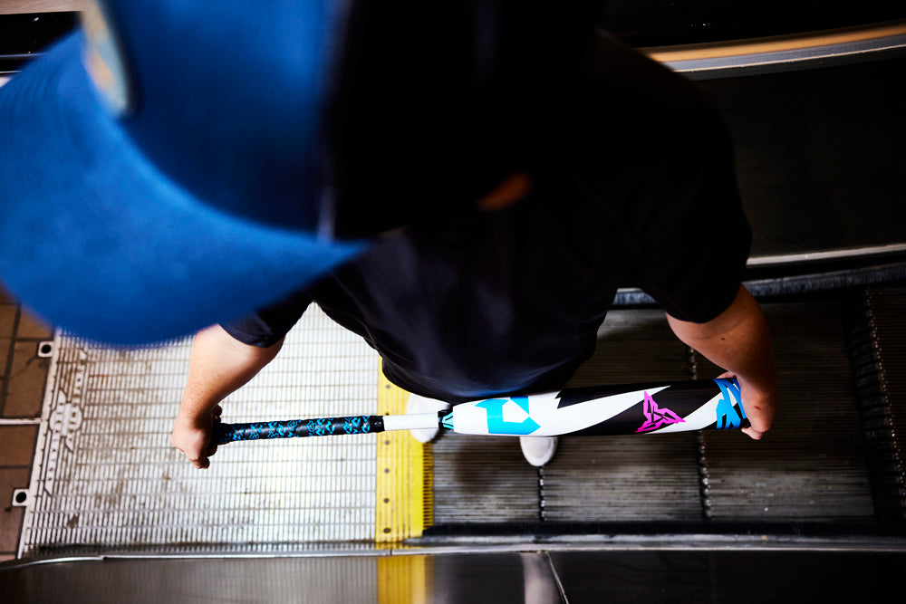 A person in a blue cap stands on an escalator, holding a scooter with colorful stickers and a used 2025 DeMarini ZEN (-10) 2 3/4" USSSA Baseball Bat: WBD2532010, viewed from above.