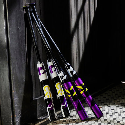 Four black, purple, and white baseball bats with yellow and white designs lean against a dark wooden wall on a tiled floor, with sunlight casting strong shadows.