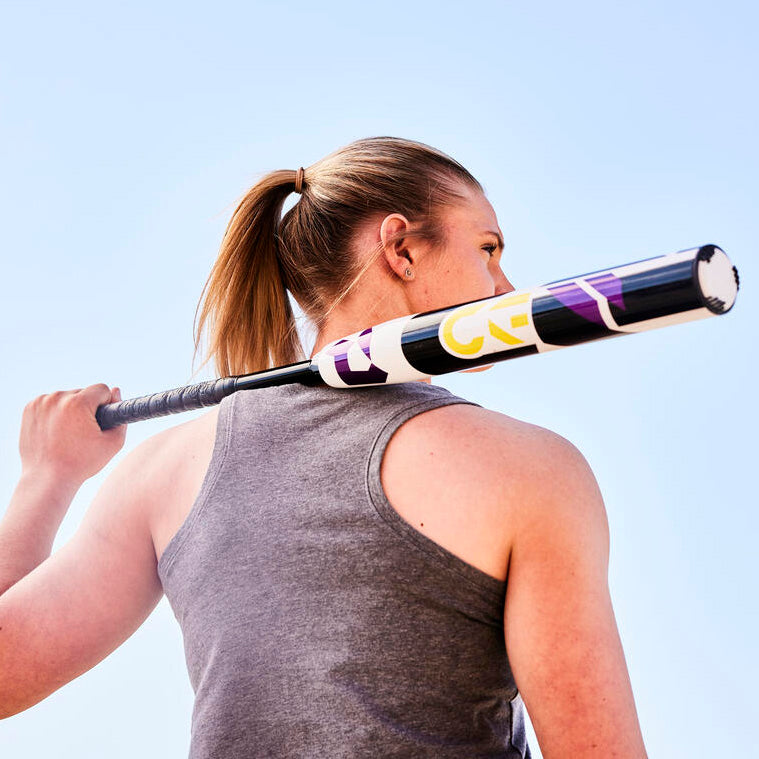 A person with a ponytail holds a baseball bat over their shoulder, facing away from the camera and wearing a gray sleeveless shirt against a bright blue sky.