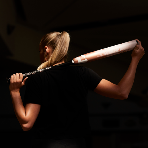 A woman with blonde hair in a ponytail stands with her back to the camera, holding a baseball bat across her shoulders in a dimly lit setting.
