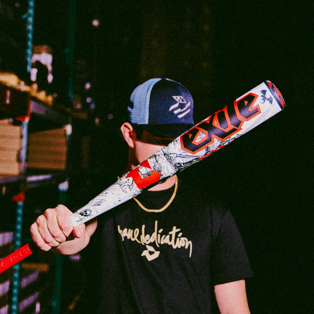 A person in a baseball cap and a "space dedication" black t-shirt holds the DeMarini 2026 Exile (-3) BBCOR Baseball Bat (WBD2589010), its one-piece alloy frame obscuring their features in a dimly lit indoor setting.