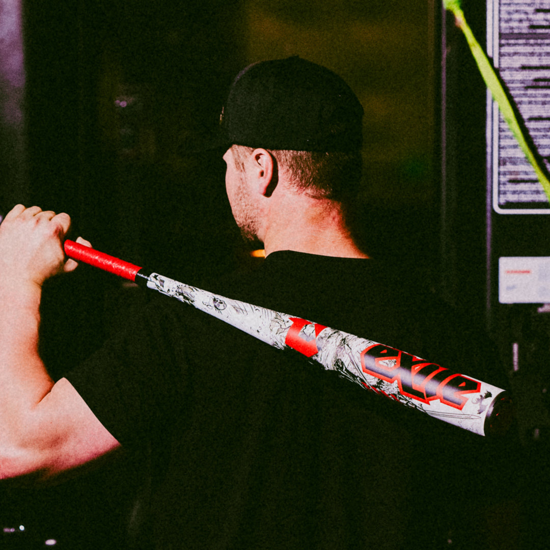 A man in a black shirt and cap stands with his back to the camera, holding a 2026 DeMarini Exile (-3) BBCOR Baseball Bat (WBD2589010) over his shoulder. The DeMarini bat stands out against the dark, blurred background.